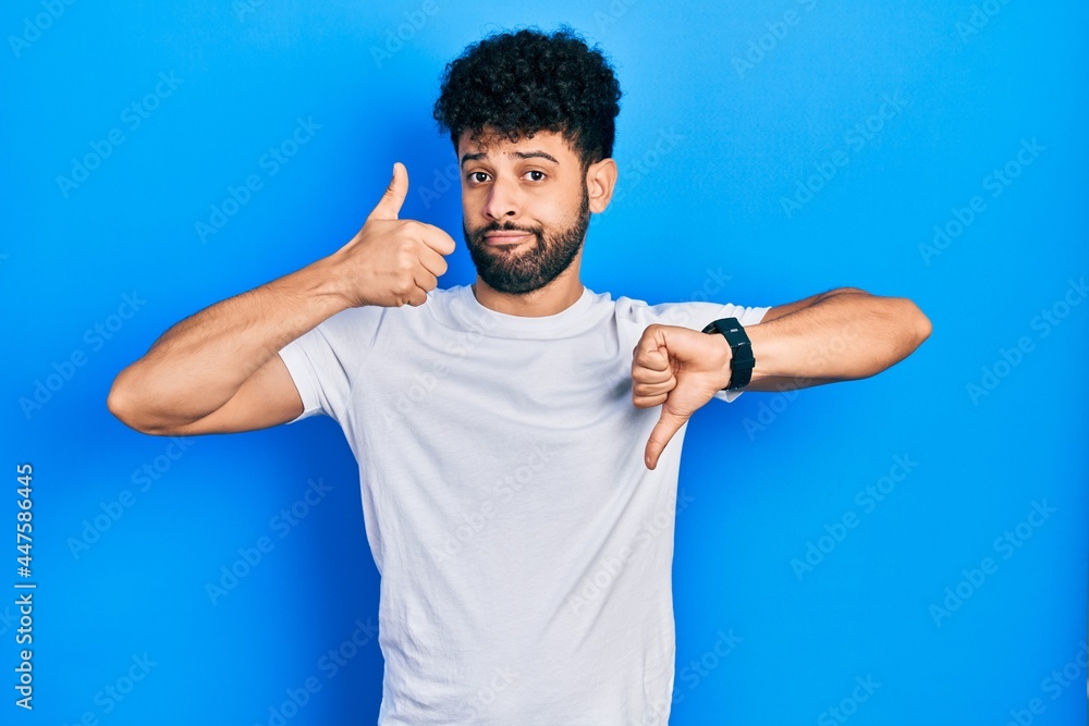 Young arab man with beard wearing casual white t shirt doing thumbs up and down, disagreement and agreement expression. crazy conflict