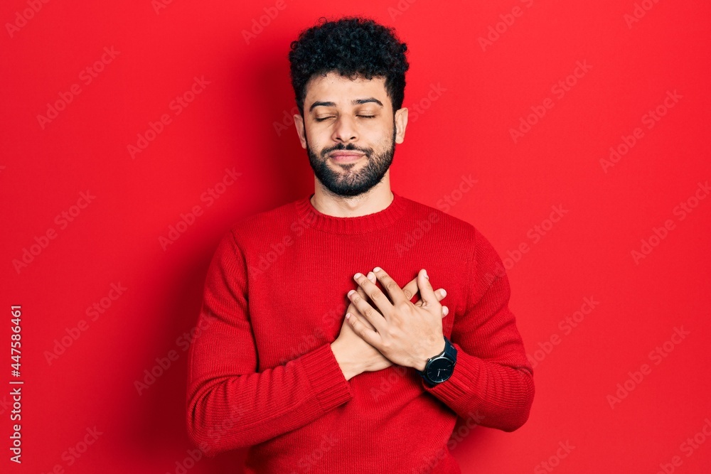 Young arab man with beard wearing casual red sweater smiling with hands on chest with closed eyes and grateful gesture on face. health concept. Young arab man with beard wearing casual red sweater smiling with hands on chest with closed eyes and grateful gesture on face. health concept.