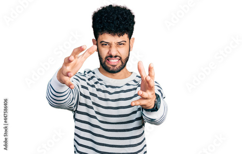 Young arab man with beard wearing casual striped sweater shouting frustrated with rage, hands trying to strangle, yelling mad