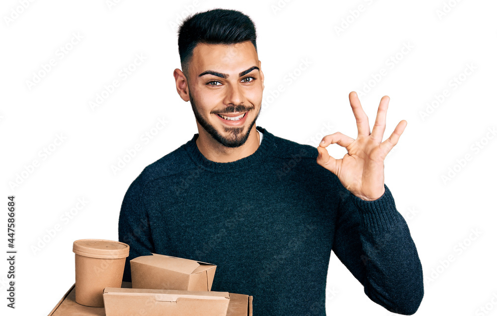 Young hispanic man with beard holding take away food doing ok sign with fingers, smiling friendly gesturing excellent symbol Young hispanic man with beard holding take away food doing ok sign with fingers, smiling friendly gesturing excellent symbol