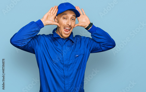 Bald man with beard wearing builder jumpsuit uniform smiling cheerful playing peek a boo with hands showing face. surprised and exited © Krakenimages.com