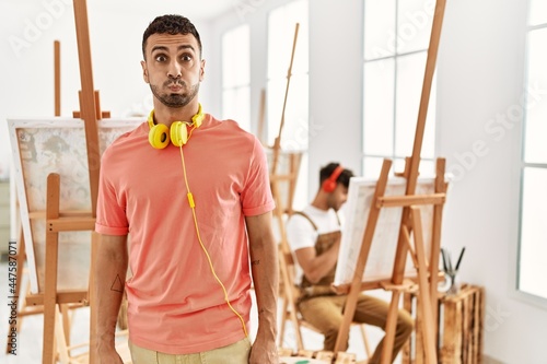 Young hispanic man at art studio puffing cheeks with funny face. mouth inflated with air, crazy expression.