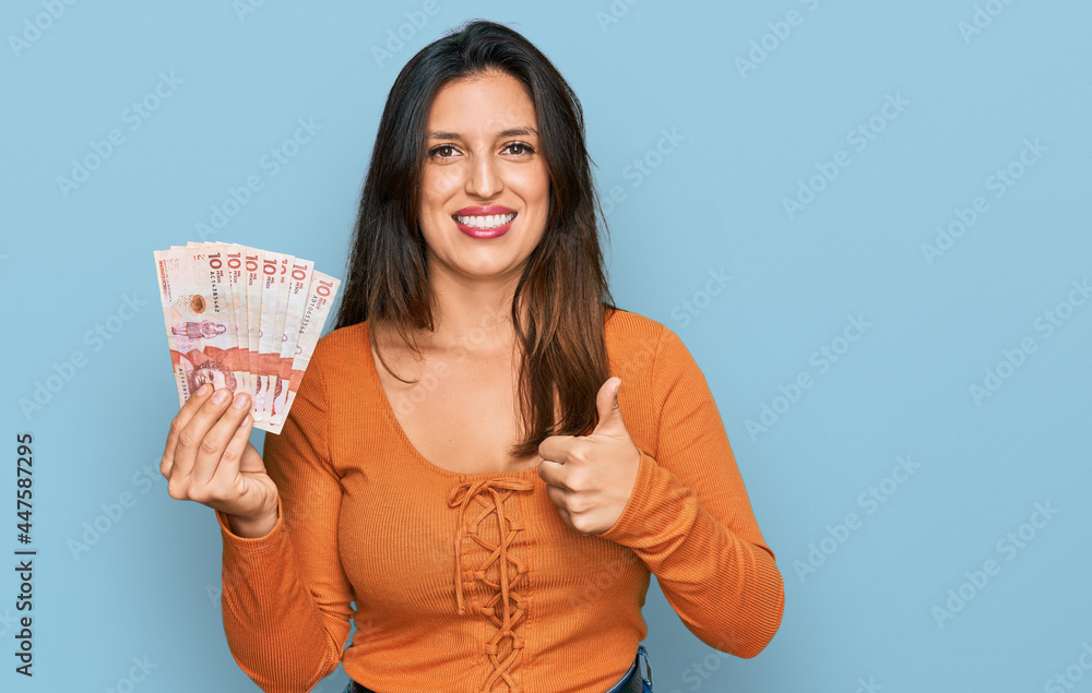 Beautiful hispanic woman holding 10 colombian pesos banknotes smiling happy and positive, thumb up doing excellent and approval sign Beautiful hispanic woman holding 10 colombian pesos banknotes smiling happy and positive, thumb up doing excellent and approval sign