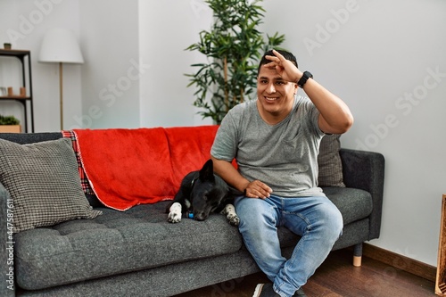 Young latin man and dog sitting on the sofa at home very happy and smiling looking far away with hand over head. searching concept. © Krakenimages.com