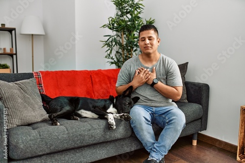 Young latin man and dog sitting on the sofa at home smiling with hands on chest with closed eyes and grateful gesture on face. health concept. © Krakenimages.com
