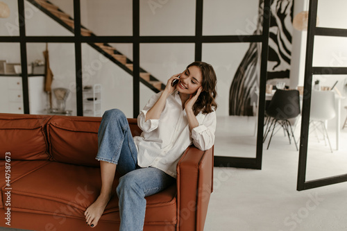 Joyful curly brunette woman in oversized white shirt and jeans sits on soft brown leathers coach and talks on phone in living room.