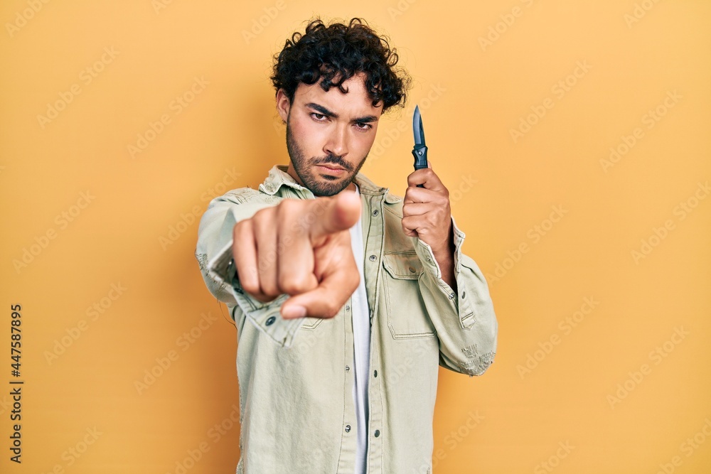 Young hispanic man holding pocket knife pointing with finger to the camera and to you, confident gesture looking serious Young hispanic man holding pocket knife pointing with finger to the camera and to you, confident gesture looking serious