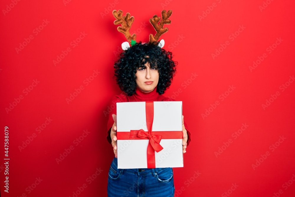 Young middle east woman wearing deer christmas hat holding gift skeptic and nervous, frowning upset because of problem. negative person. Young middle east woman wearing deer christmas hat holding gift skeptic and nervous, frowning upset because of problem. negative person.