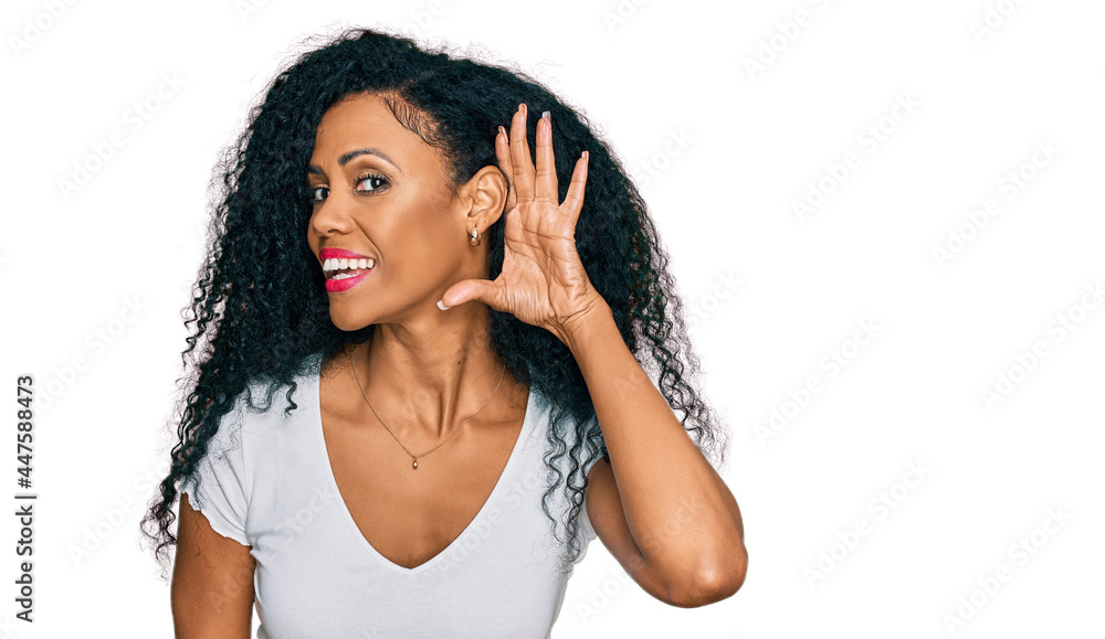 Middle age african american woman wearing casual white t shirt smiling with hand over ear listening an hearing to rumor or gossip. deafness concept. Middle age african american woman wearing casual white t shirt smiling with hand over ear listening an hearing to rumor or gossip. deafness concept.