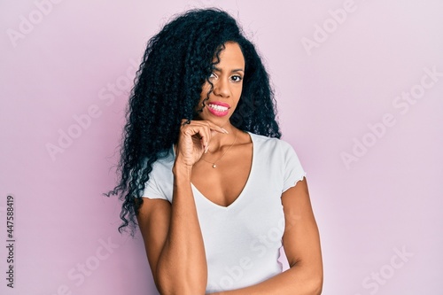 Middle age african american woman wearing casual white t shirt smiling looking confident at the camera with crossed arms and hand on chin. thinking positive.