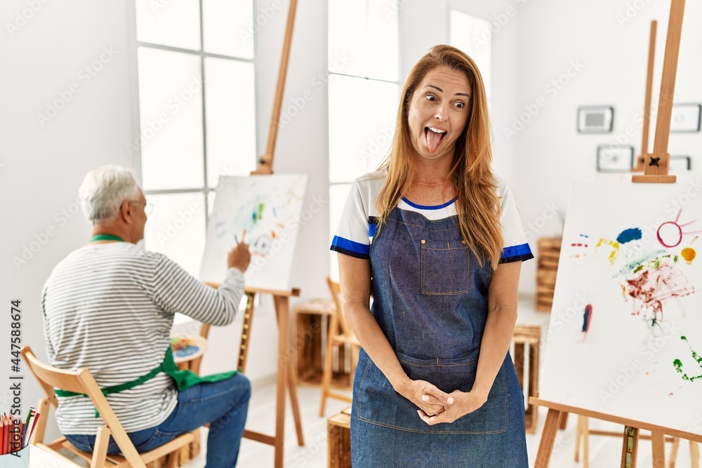 Hispanic woman wearing apron at art studio sticking tongue out happy with funny expression. emotion concept.