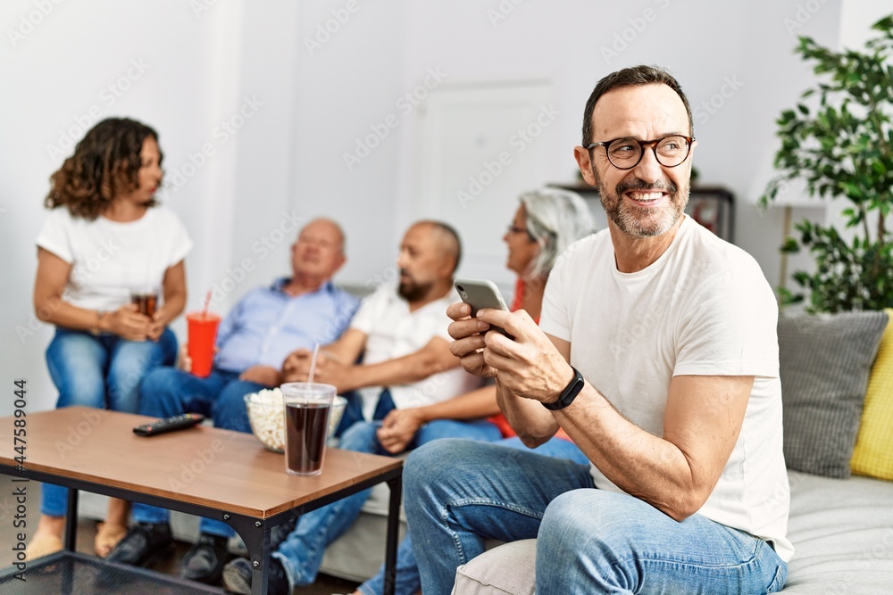 Fototapeta premium Group of middle age friends sitting on the sofa speaking. Man smiling happy using smartphone at home.