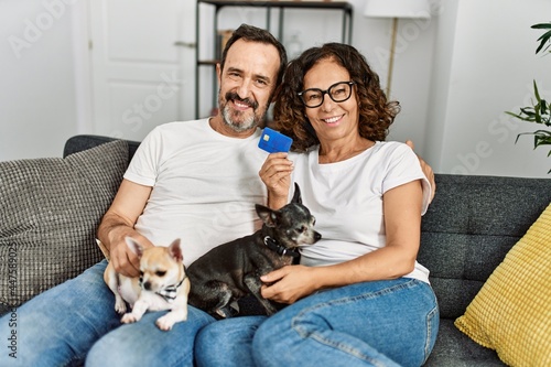 Middle age hispanic couple smiling happy and holding credit card. Sitting on the sofa with dogs at home.