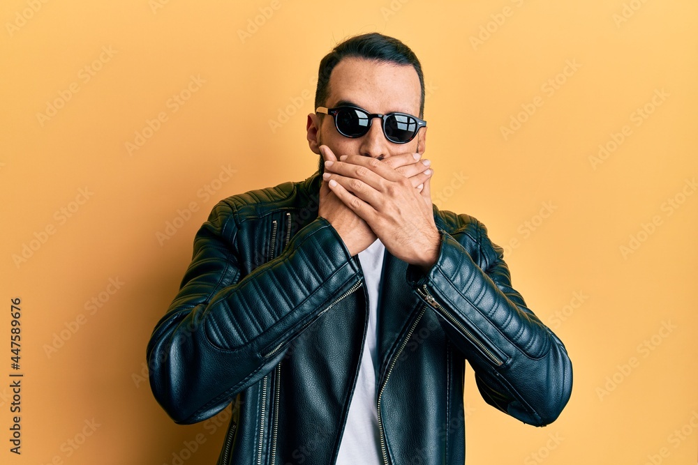 Obraz premium Young man with beard wearing black leather jacket and sunglasses shocked covering mouth with hands for mistake. secret concept.