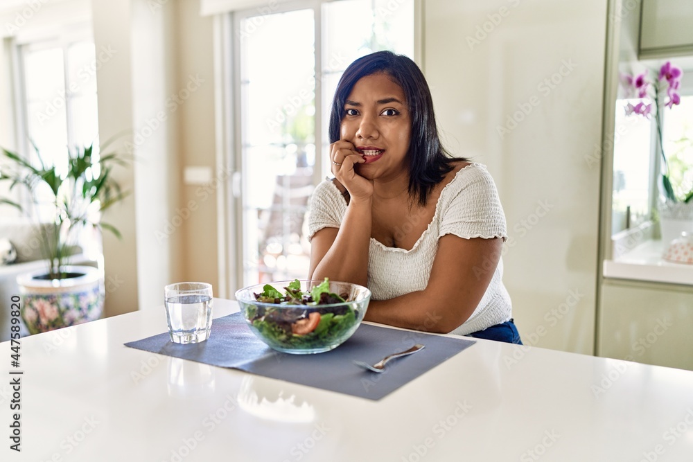 Young hispanic woman eating healthy salad at home looking stressed and nervous with hands on mouth biting nails. anxiety problem.