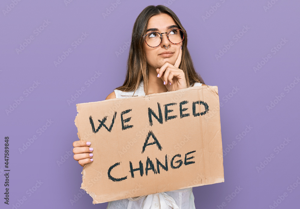 Young hispanic woman holding we need a change banner serious face thinking about question with hand on chin, thoughtful about confusing idea Young hispanic woman holding we need a change banner serious face thinking about question with hand on chin, thoughtful about confusing idea