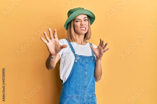 Young caucasian blonde woman wearing denim jumpsuit and hat with 90s style disgusted expression, displeased and fearful doing disgust face because aversion reaction. with hands raised