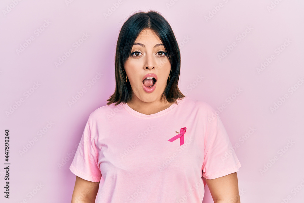 Fototapeta premium Young hispanic woman wearing pink cancer ribbon on t shirt in shock face, looking skeptical and sarcastic, surprised with open mouth