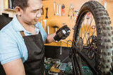 male bicycle mechanic in apron holding bottle while applying lubricant spray to bicycle chain in workshop