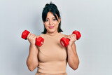Young hispanic woman wearing sportswear using dumbbells relaxed with serious expression on face. simple and natural looking at the camera.