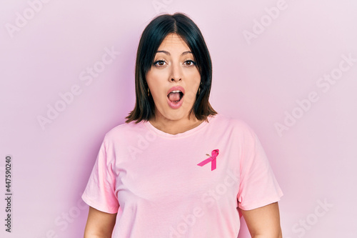 Young hispanic woman wearing pink cancer ribbon on t shirt in shock face, looking skeptical and sarcastic, surprised with open mouth