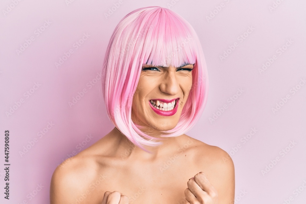 Young man wearing woman make up wearing pink wig excited for success with arms raised and eyes closed celebrating victory smiling. winner concept. Young man wearing woman make up wearing pink wig excited for success with arms raised and eyes closed celebrating victory smiling. winner concept.