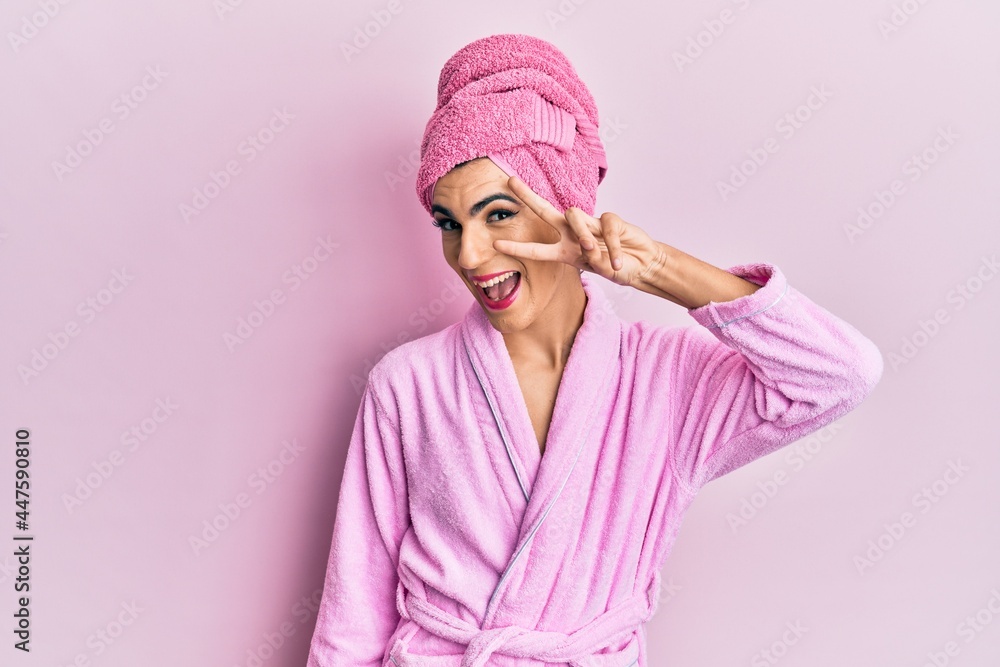 Young man wearing woman make up wearing shower towel on head and bathrobe doing peace symbol with fingers over face, smiling cheerful showing victory Young man wearing woman make up wearing shower towel on head and bathrobe doing peace symbol with fingers over face, smiling cheerful showing victory