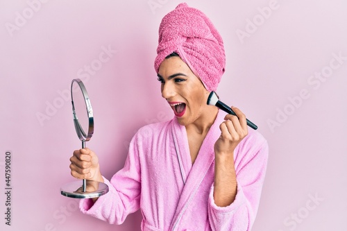 Young man wearing woman make up wearing pink robe applying blush celebrating crazy and amazed for success with open eyes screaming excited. photo