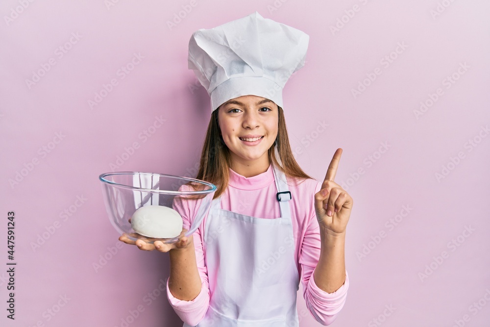Beautiful brunette little girl wearing chef hat holding bread dough smiling with an idea or question pointing finger with happy face, number one Beautiful brunette little girl wearing chef hat holding bread dough smiling with an idea or question pointing finger with happy face, number one