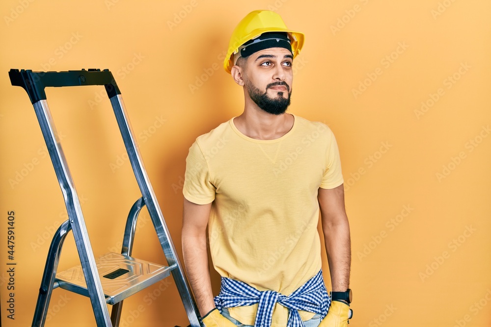 Handsome man with beard by construction stairs wearing hardhat smiling looking to the side and staring away thinking. Handsome man with beard by construction stairs wearing hardhat smiling looking to the side and staring away thinking.