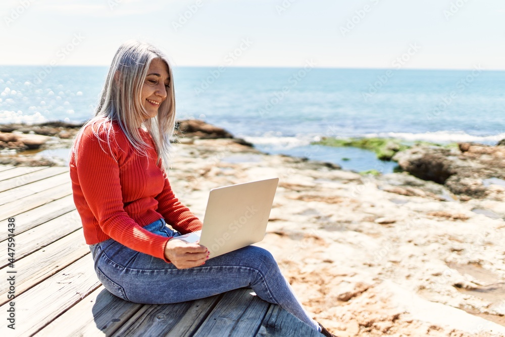 Middle age grey-haired woman using laptop sitting on the bench at the beach.