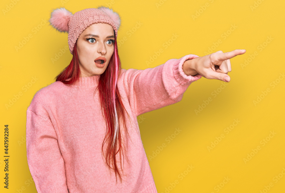 Young caucasian woman wearing casual clothes and wool cap pointing with finger surprised ahead, open mouth amazed expression, something on the front Young caucasian woman wearing casual clothes and wool cap pointing with finger surprised ahead, open mouth amazed expression, something on the front
