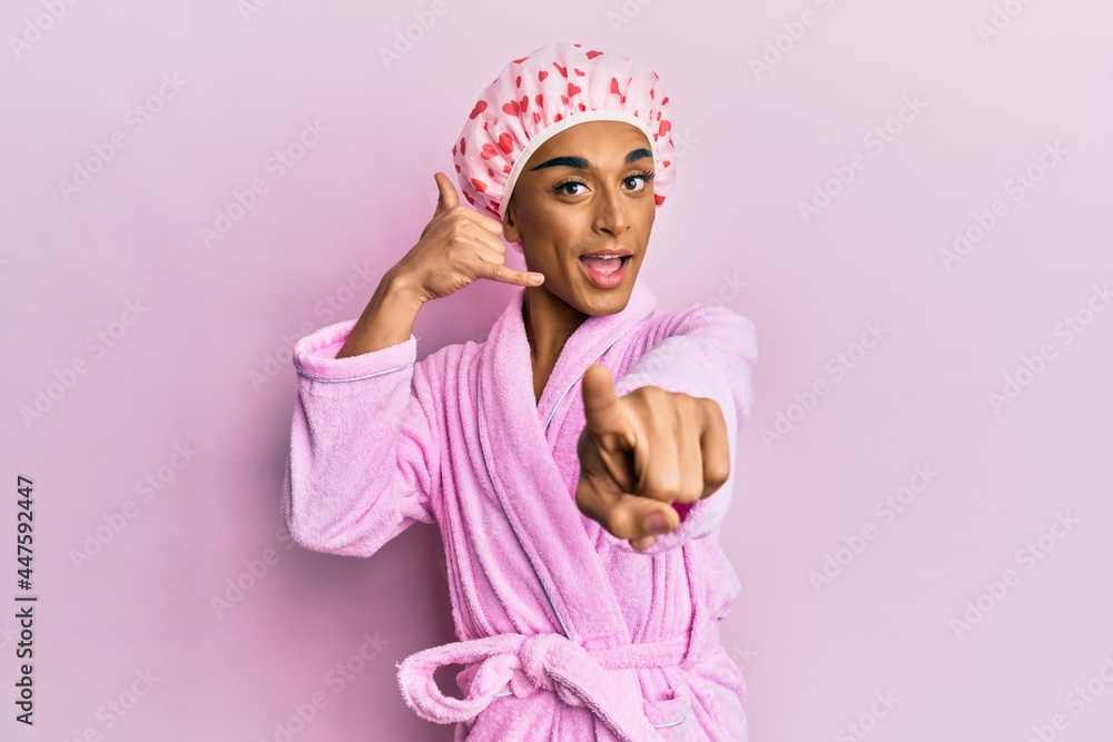 Hispanic man wearing make up wearing shower towel cap and bathrobe smiling doing talking on the telephone gesture and pointing to you. call me.