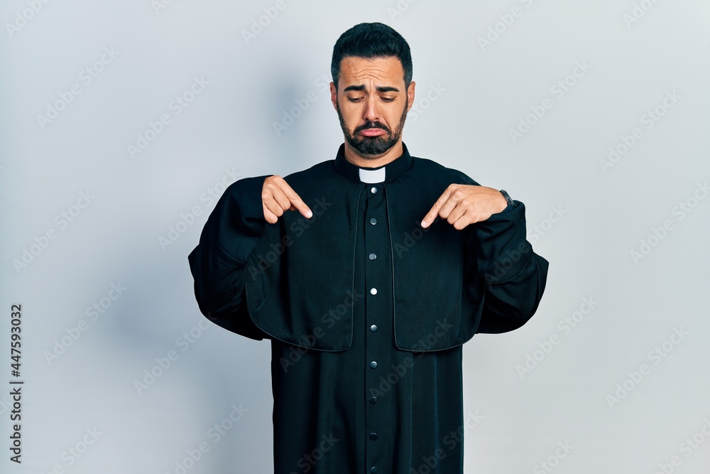 Handsome hispanic man with beard wearing catholic priest robe pointing down looking sad and upset, indicating direction with fingers, unhappy and depressed. Handsome hispanic man with beard wearing catholic priest robe pointing down looking sad and upset, indicating direction with fingers, unhappy and depressed.