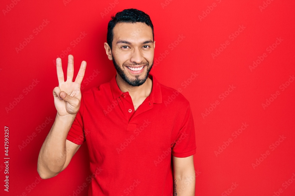 Naklejka premium Hispanic man with beard wearing casual red t shirt showing and pointing up with fingers number three while smiling confident and happy.