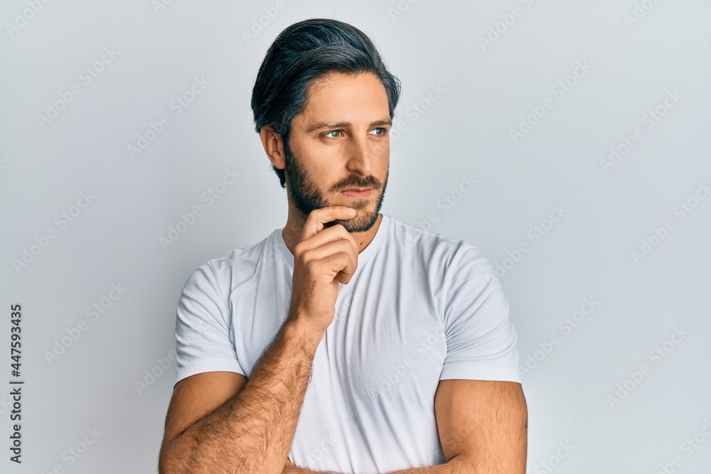 Young hispanic man wearing casual white t shirt thinking concentrated about doubt with finger on chin and looking up wondering Young hispanic man wearing casual white t shirt thinking concentrated about doubt with finger on chin and looking up wondering