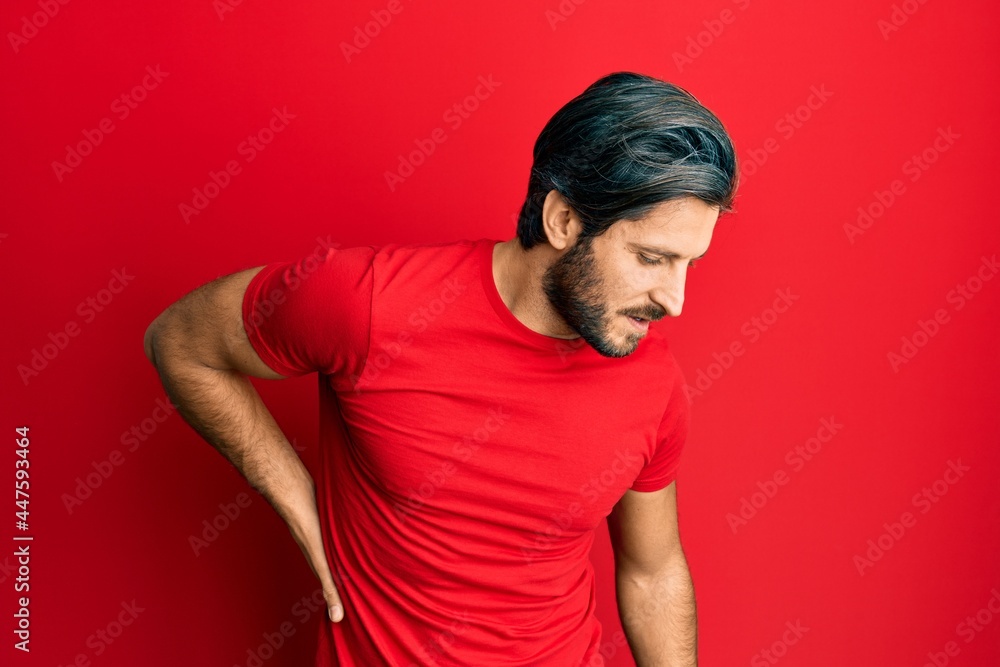 Young hispanic man wearing casual red t shirt suffering of backache, touching back with hand, muscular pain Young hispanic man wearing casual red t shirt suffering of backache, touching back with hand, muscular pain