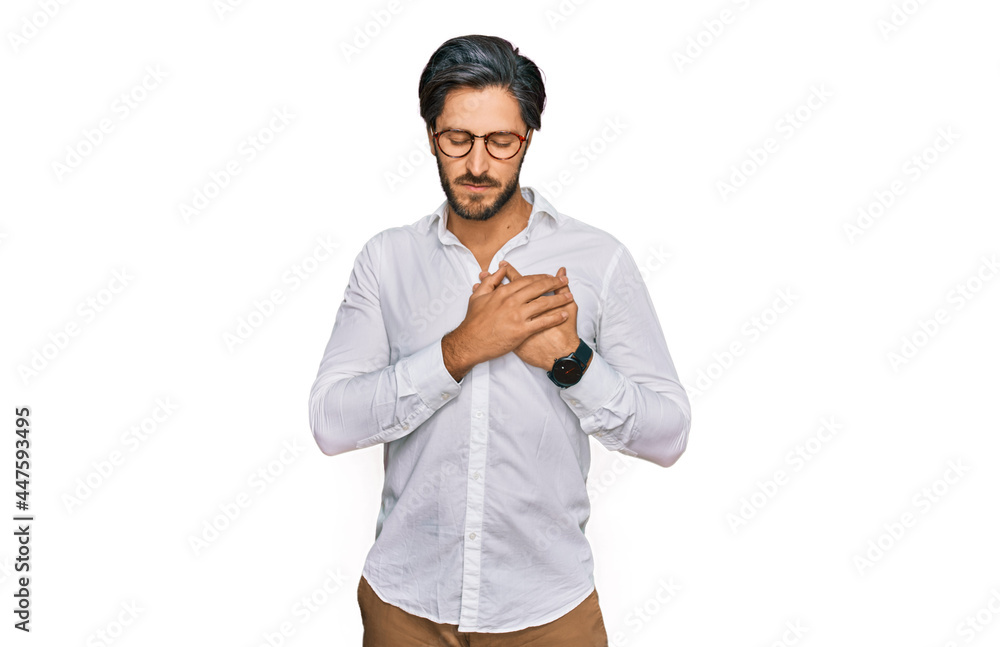 Young hispanic man wearing business shirt and glasses smiling with hands on chest with closed eyes and grateful gesture on face. health concept. Young hispanic man wearing business shirt and glasses smiling with hands on chest with closed eyes and grateful gesture on face. health concept.