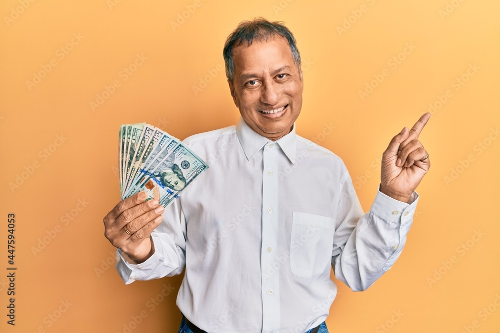 Middle age indian man holding dollars smiling happy pointing with hand and finger to the side Middle age indian man holding dollars smiling happy pointing with hand and finger to the side