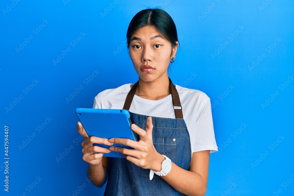 Young chinese woman wearing cook apron using touchpad clueless and confused expression. doubt concept. Young chinese woman wearing cook apron using touchpad clueless and confused expression. doubt concept.