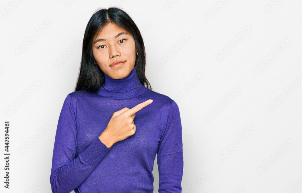 Beautiful young asian woman wearing casual clothes pointing with hand finger to the side showing advertisement, serious and calm face Beautiful young asian woman wearing casual clothes pointing with hand finger to the side showing advertisement, serious and calm face