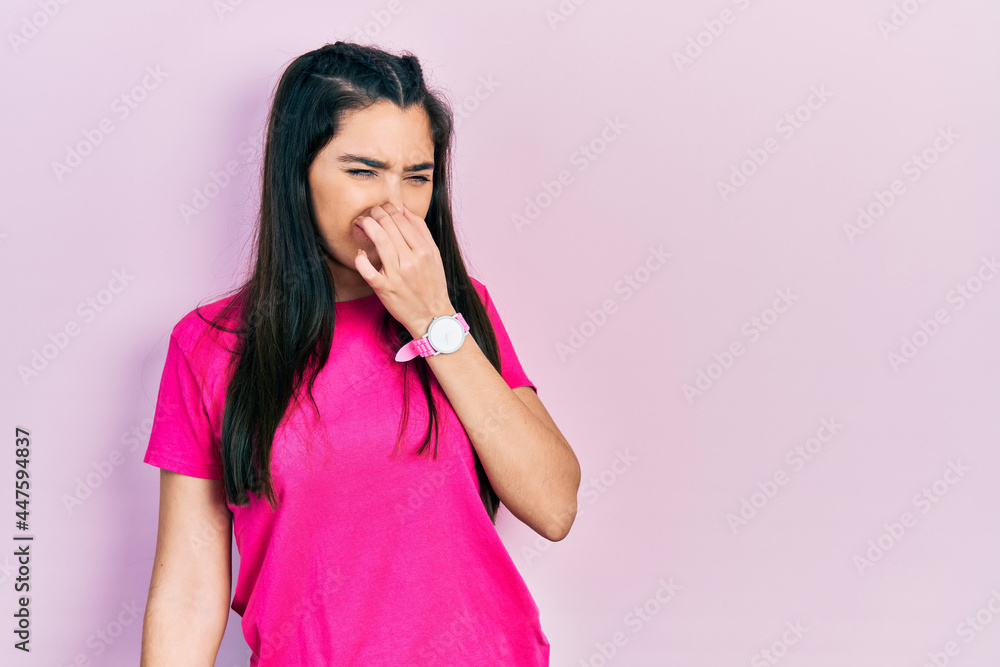 Young hispanic girl wearing casual pink t shirt smelling something stinky and disgusting, intolerable smell, holding breath with fingers on nose. bad smell Young hispanic girl wearing casual pink t shirt smelling something stinky and disgusting, intolerable smell, holding breath with fingers on nose. bad smell