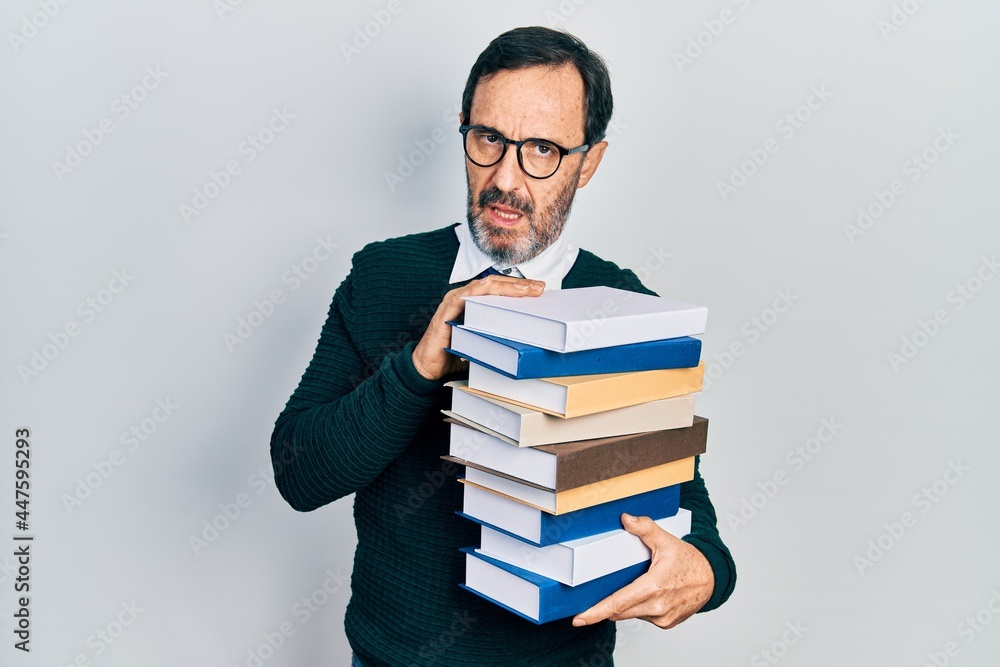 Middle age hispanic man holding a pile of books in shock face, looking skeptical and sarcastic, surprised with open mouth Middle age hispanic man holding a pile of books in shock face, looking skeptical and sarcastic, surprised with open mouth