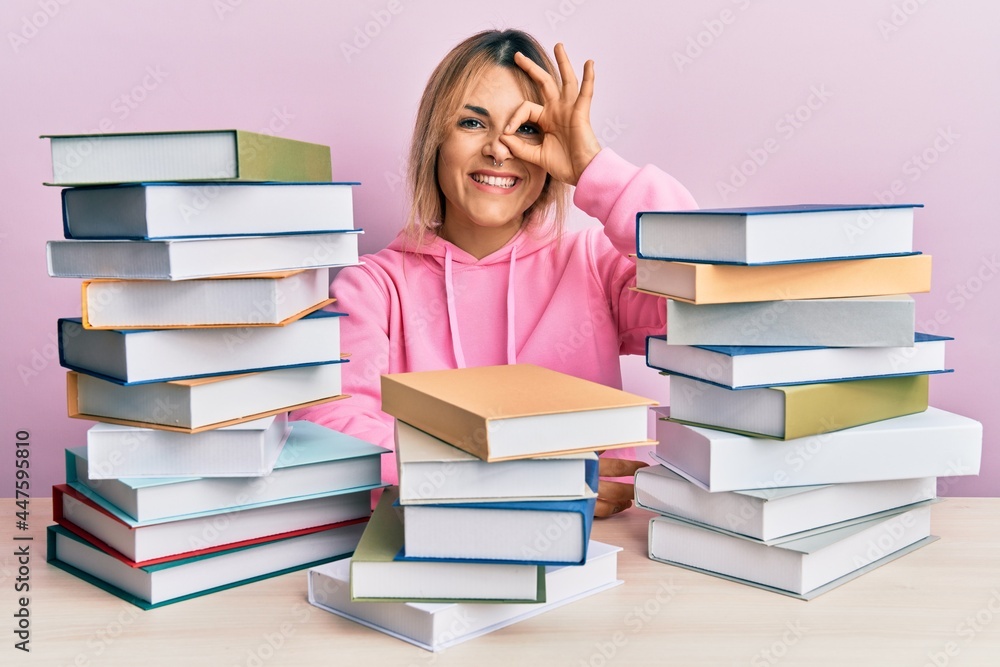 Young caucasian woman sitting on the table with books doing ok gesture with hand smiling, eye looking through fingers with happy face. Young caucasian woman sitting on the table with books doing ok gesture with hand smiling, eye looking through fingers with happy face.