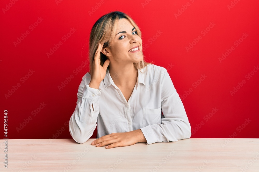 Young caucasian woman wearing casual clothes sitting on the table smiling with hand over ear listening an hearing to rumor or gossip. deafness concept. Young caucasian woman wearing casual clothes sitting on the table smiling with hand over ear listening an hearing to rumor or gossip. deafness concept.
