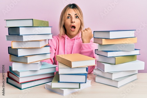 Young caucasian woman sitting on the table with books surprised pointing with hand finger to the side, open mouth amazed expression.