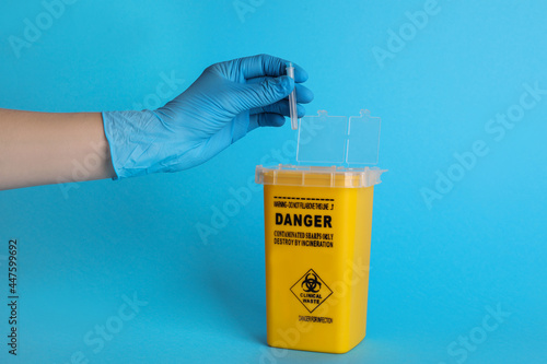 Doctor throwing used syringe needle into sharps container on light blue background, closeup photo