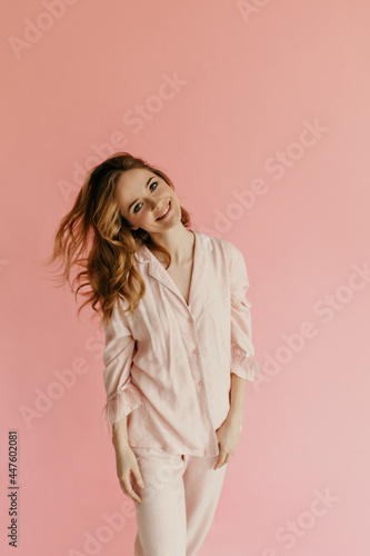 Charming cheerful woman with curly modern hairstyle in pink night-suit looking into camera and smiling on isolated background..