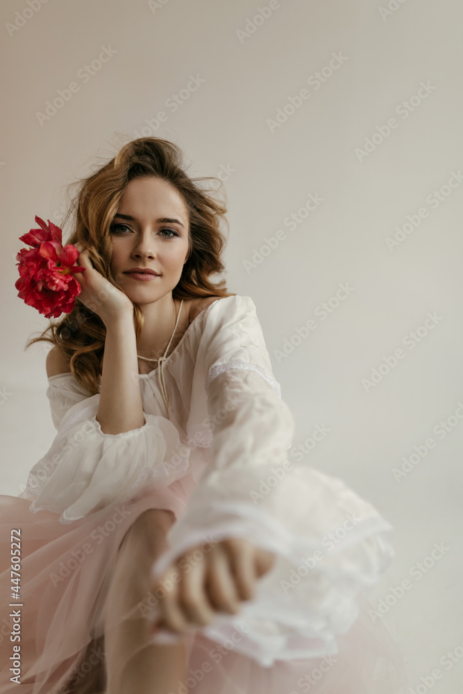 Stylish lovely girl with curly hairstyle in shirt with long sleeves and pink skirt posing and holding red peony on white background.. Stylish lovely girl with curly hairstyle in shirt with long sleeves and pink skirt posing and holding red peony on white background..