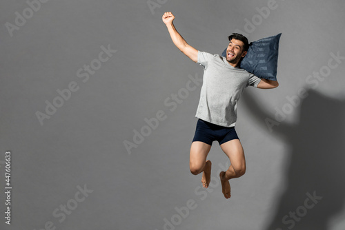 Fresh and energetic young Caucasian man in sleepwear jumping with pillow after wake up in light gray isolated background, good sleep hygiene concept